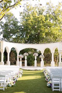 An outdoor wedding venue with white chairs arranged in rows, floral decorations along the aisle, string lights overhead, and a white archway in front, surrounded by trees.