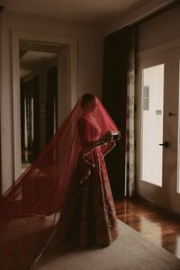 South Asian bride in embroidered red lehenga and flowing dupatta stands poised in warm moody indoor light before her ceremony