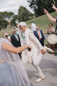Indian groom in ivory sherwani and feathered turban dances joyfully during baraat procession with dhol drummer and guests outdoors