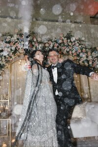 Couple celebrating with arms raised amid bursts of CO2 cannon fog at their reception, the bride in a sparkling silver outfit and the groom in a black suit, beneath a floral arch at a Hyatt Chesapeake Bay Sikh wedding.