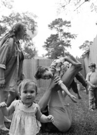 A father lifts his son up by his legs while the mother looks over at them and his baby sister gazes right at the camera during their at home family photo session in houston texas
