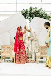 Smiling bride in red gold lehenga places a floral garland on her groom during an elegant indoor Indian wedding ceremony