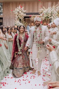Radiant Indian bride and groom walk joyfully down the aisle as guests shower them with red rose petals
