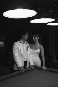 Bride and groom share a smiling glance leaning against a pool table beneath dramatic pendant lights at Belmont Country Club
