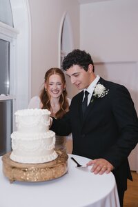 Laughing bride and smiling groom cut their white tiered wedding cake together inside an elegant chapel reception venue