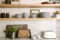 Warm farmhouse kitchen with open wood shelves, stacked white plates, framed landscape art, greenery, vintage cutting boards, and a white toaster.