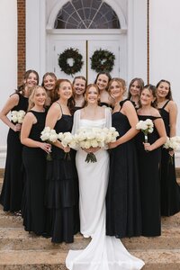 Radiant bride in white gown poses with ten smiling bridesmaids in black dresses holding white roses outside a classic chapel