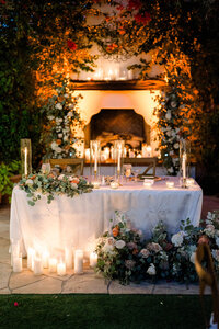 Candlelit wedding sweetheart table surrounded by white and peach flowers and greenery at an outdoor reception.