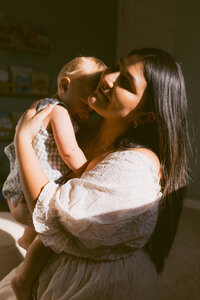 Mother embraces her baby in the sunshine of their home during their at home family photo session near houston texas