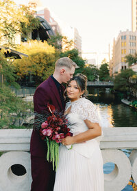 Bride and groom embracing along the San Antonio River Walk with downtown skyline and fall colors during their wedding portraits.