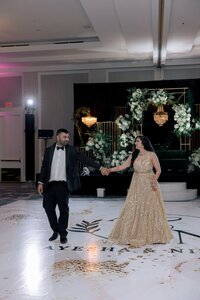 Bride in a gold sequined gown holds hands with her tuxedoed groom during their first dance on a custom monogrammed ballroom floor