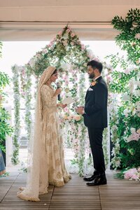 A bride in gold-embroidered attire reads vows to her groom beneath a lush pink-and-white floral arch at an intimate Muslim wedding ceremony