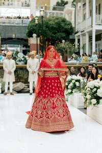 Radiant Indian bride in gold-embroidered red lehenga walks smiling down the aisle at an outdoor garden ceremony