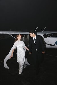 Laughing bride in a flowing white cape gown and groom in a black suit walk hand-in-hand beside a small aircraft on a dark airfield