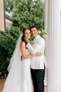 Smiling bride and groom embrace beside white columns at Belmont Country Club amid lush greenery on their wedding day
