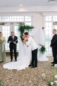 Bride and groom share a passionate first kiss beneath a lush greenery archway in a bright, elegant country club ballroom
