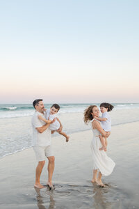 A mom, dad, and two toddler boys playing and laughing on the beach during their sunset family photography session at Wrightsville Beach, North Carolina