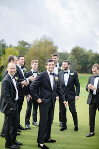 Groom laughs with his groomsmen in black tuxedos on a grassy outdoor law