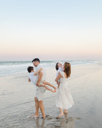 A mom, dad, and two toddler boys playing and laughing on the beach during their sunset family photography session at Wrightsville Beach, North Carolina