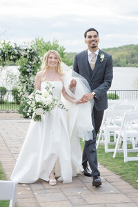 Newlyweds walk hand in hand from their lakeside ceremony beneath a white floral arch