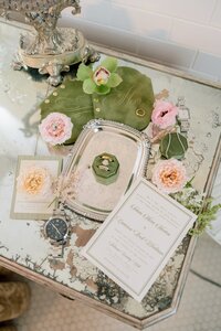 Elegant wedding flat lay featuring invitation suite, diamond rings in a green velvet box, pink roses, and watches on an antique mirrored vanity