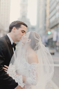 Bride and groom kiss under a bow-topped veil on a city street with traffic lights behind them