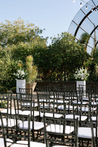 Outdoor wedding ceremony setup with black chairs, white floral arrangements and lush desert garden landscaping.