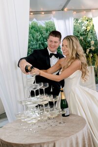 Smiling bride and groom together pouring champagne over a crystal coupe tower at an elegant tented wedding reception