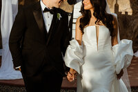 Bride in a modern white gown with sculptural sleeves holding hands with groom in a black tuxedo.