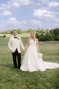 Bride and groom hold hands in a countryside field with fences and rolling hills behind them