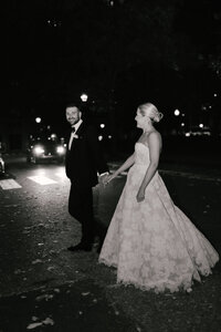 Bride and groom walk hand in hand down a city street lit by car headlights at night
