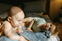 A toddler lays on the bed in the sunshine beside her mother gazing at her baby sister during their at home family photo session near houston texas