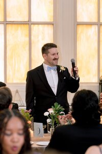 Smiling groom in black tuxedo and blue bow tie delivers a heartfelt toast holding a microphone at a warmly lit wedding reception