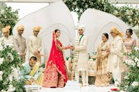 Bride in red lehenga and groom in ivory sherwani exchange rings beneath a white floral archway at their elegant Hindu ceremony