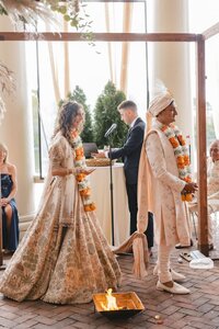 Smiling bride and groom in ornate cream and gold Indian attire circle a sacred fire beneath a wooden mandap archway