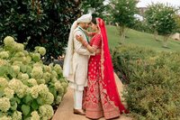 South Asian bride in red lehenga and groom in ivory sherwani share an intimate forehead-touch moment amid lush garden greenery