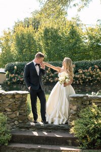 Groom in black tuxedo gallantly kisses his smiling bride's hand beside a rustic stone wall surrounded by blooming roses
