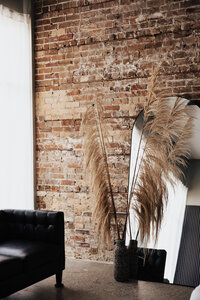 Tall pampas grass in front of a full-length mirror against exposed brick wall, with black leather couch and sheer curtain, styled detail from Love, Kay Collective’s boudoir studio in Tampa.