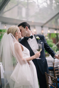 Bride and groom share a kiss while holding wine glasses at a tented reception