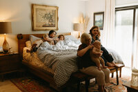 Four generations of women gather on a bed together in the sunshine during their at home family photo session near houston texas