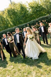 Bride and groom hold hands laughing while wedding party in black tuxedos and olive gowns walks through sunny garden