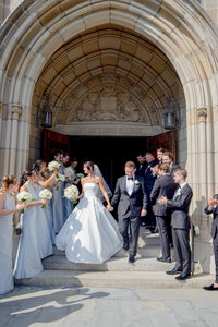 Bride and groom exit church through a stone archway as their bridal party cheers
