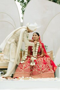 Smiling bride in red embroidered lehenga receives floral garland from groom in ivory sherwani beneath elegant white archway