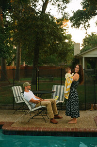 A film photo of a mother and father sitting beside their pool while the mother holds her baby during their at home family photo session near houston texas