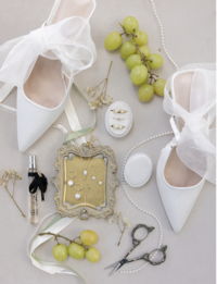Elegant wedding flat lay with white bridal heels, a vintage gold tray with jewelry, grapes, perfume, and a string of pearls on a soft background.
