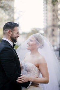 Bride and groom smile at each other on a city street with her veil catching the light