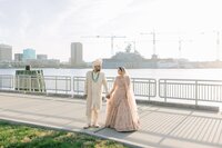 South Asian bride in blush lehenga and groom in ivory sherwani hold hands walking along a sunlit Norfolk harbor promenade