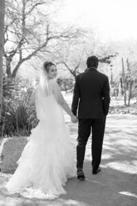 Black-and-white wedding portrait of a bride looking back while holding hands with the groom along a desert garden path.