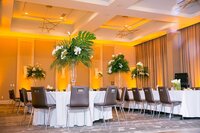 Tables with white tablecloths, brown wooden chairs and tall floral arrangements with lush greenery, palm fronds, roses and orchids