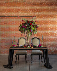 Wedding sweetheart table styled with a dramatic red and purple floral arrangement, black linens, candles and vintage chairs.